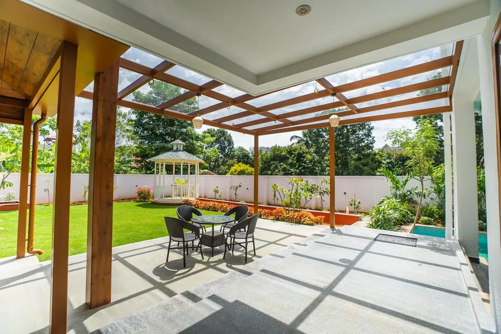 Brown metal grid and clear glass roof above a paved patio with round dining table, beside a lawn.