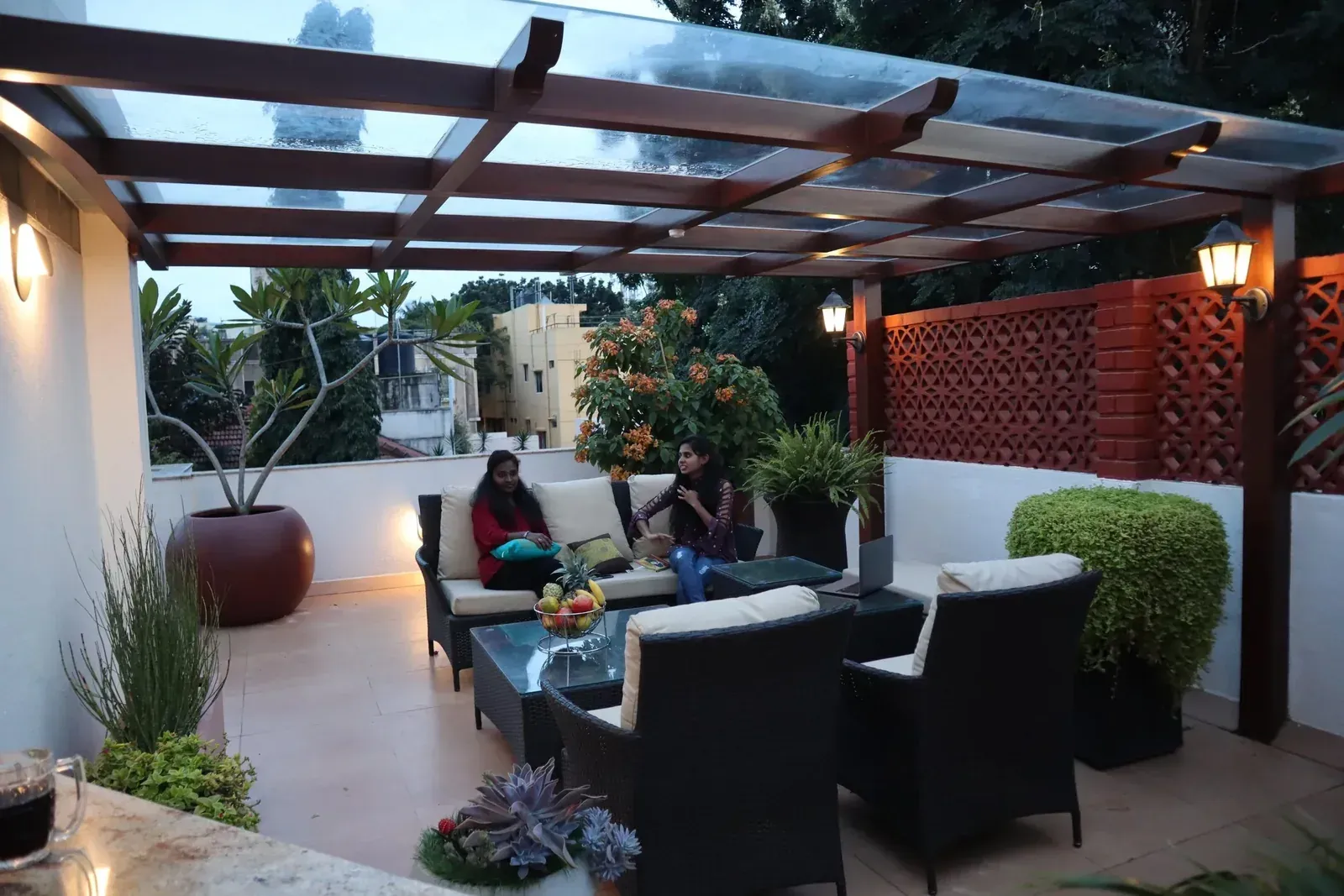 Brown-framed clear glass roof over rooftop lounge seating at dusk, beside red lattice brickwork.