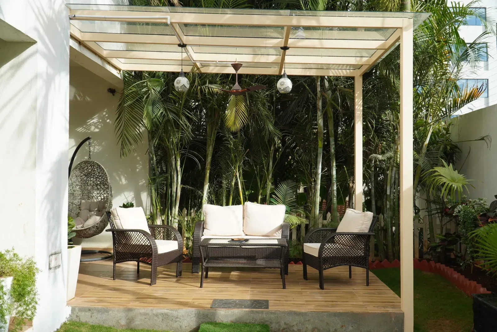 Cream metal frame and clear glass roof over woven outdoor seating, with hanging lights and a ceiling fan.