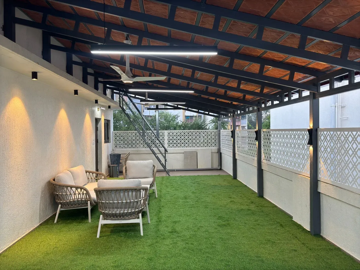 Long view of the lit terrace with seating, green flooring, staircase and patterned fencing
