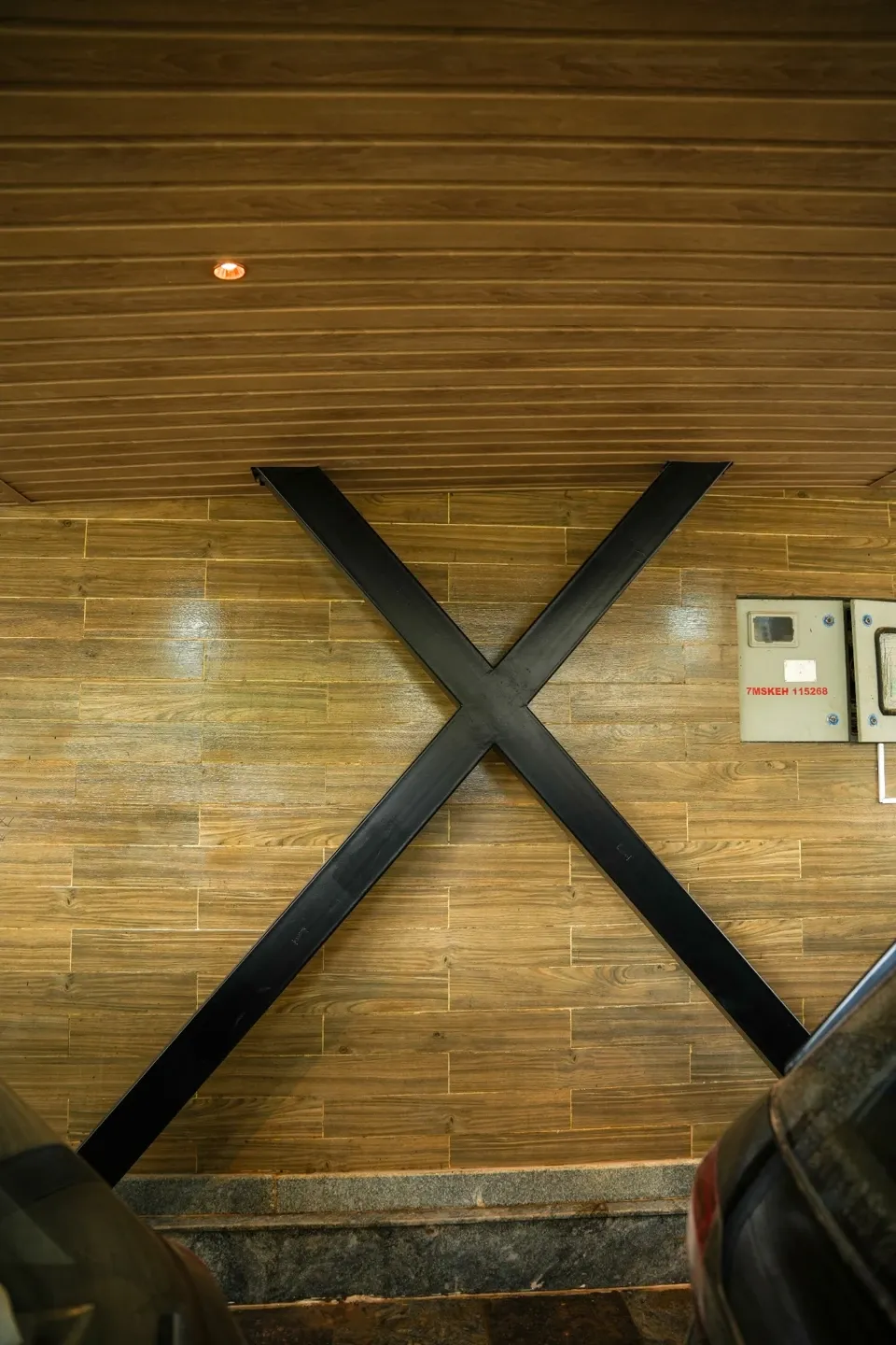 Portrait photograph of a dark X-shaped metal support against a timber-toned wall below the canopy
