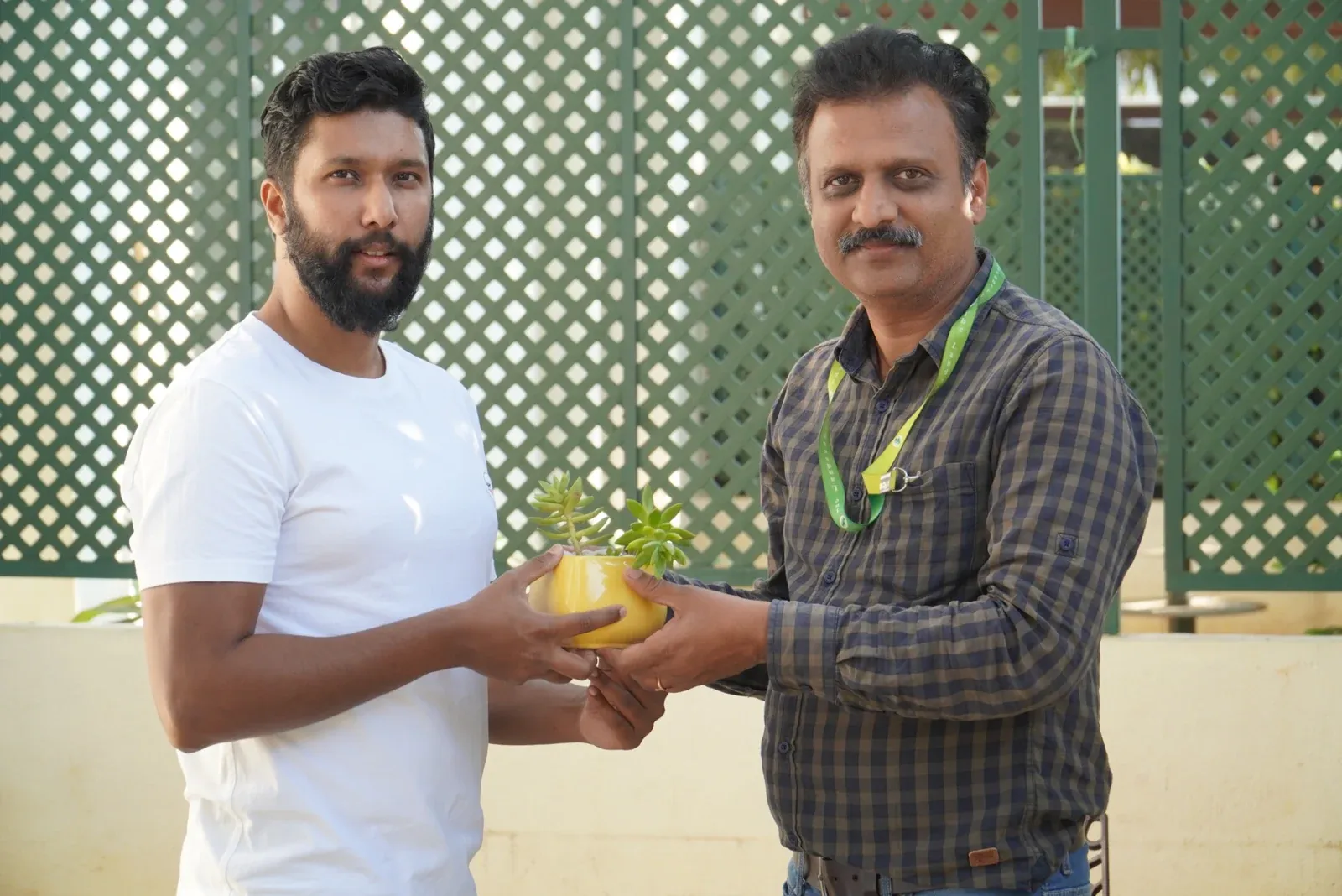Two people holding a potted plant beside a lattice privacy screen